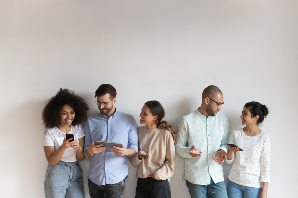 Group of smiling young mixed race people standing near white wall in row with different gadgets