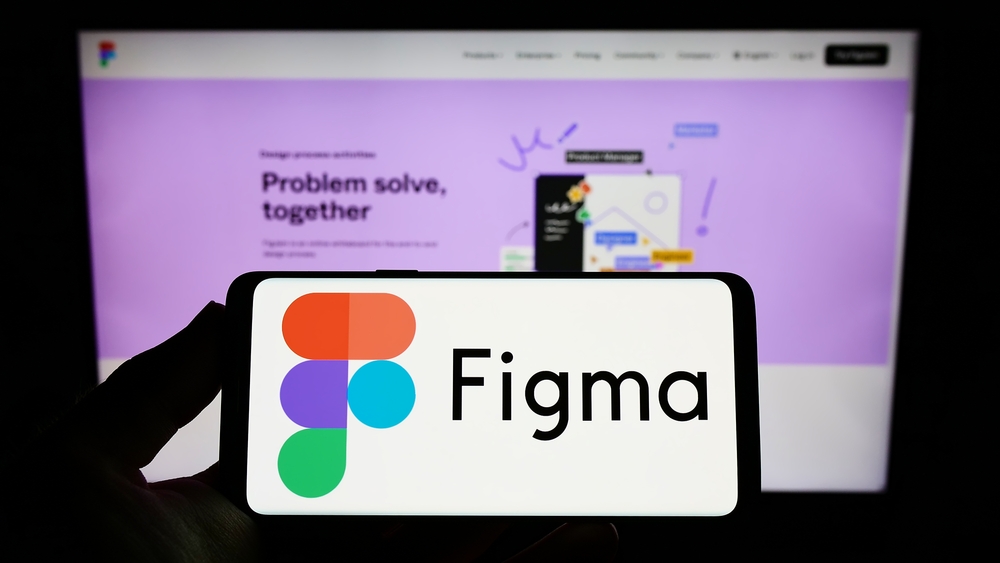 Person holding smartphone with logo of Figma Inc. on mobile screen in front of desktop screen