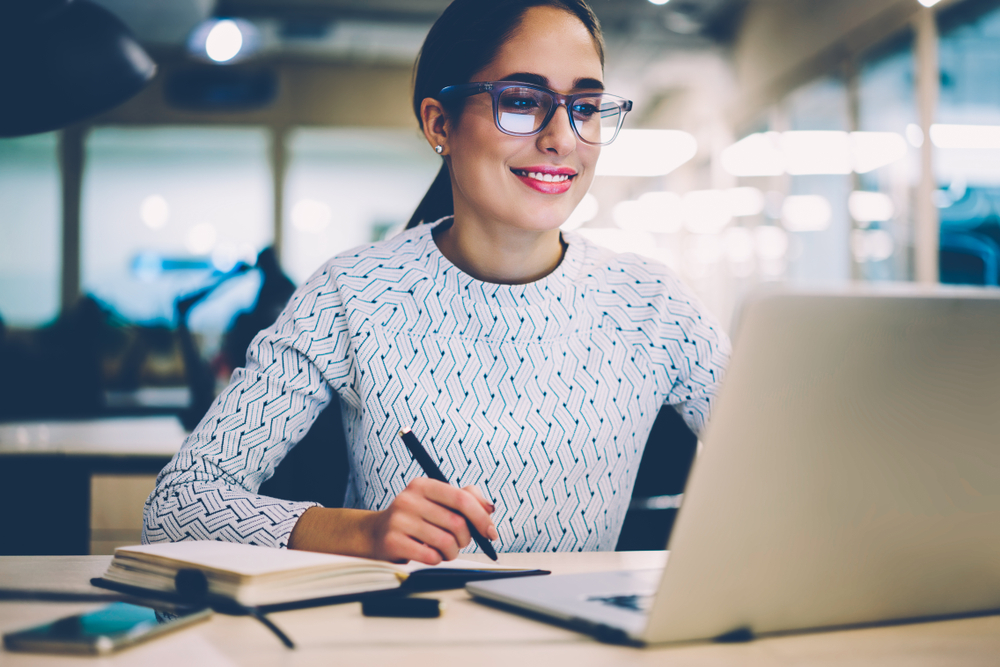 woman with glasses researching using laptop and taking down notes