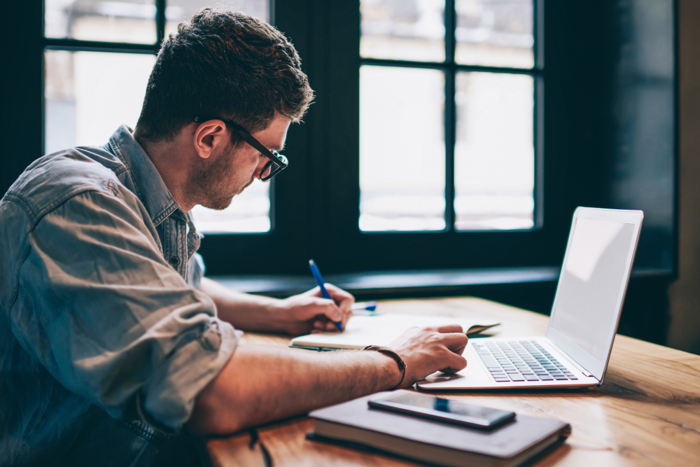 man writing on a notebook while using his laptop